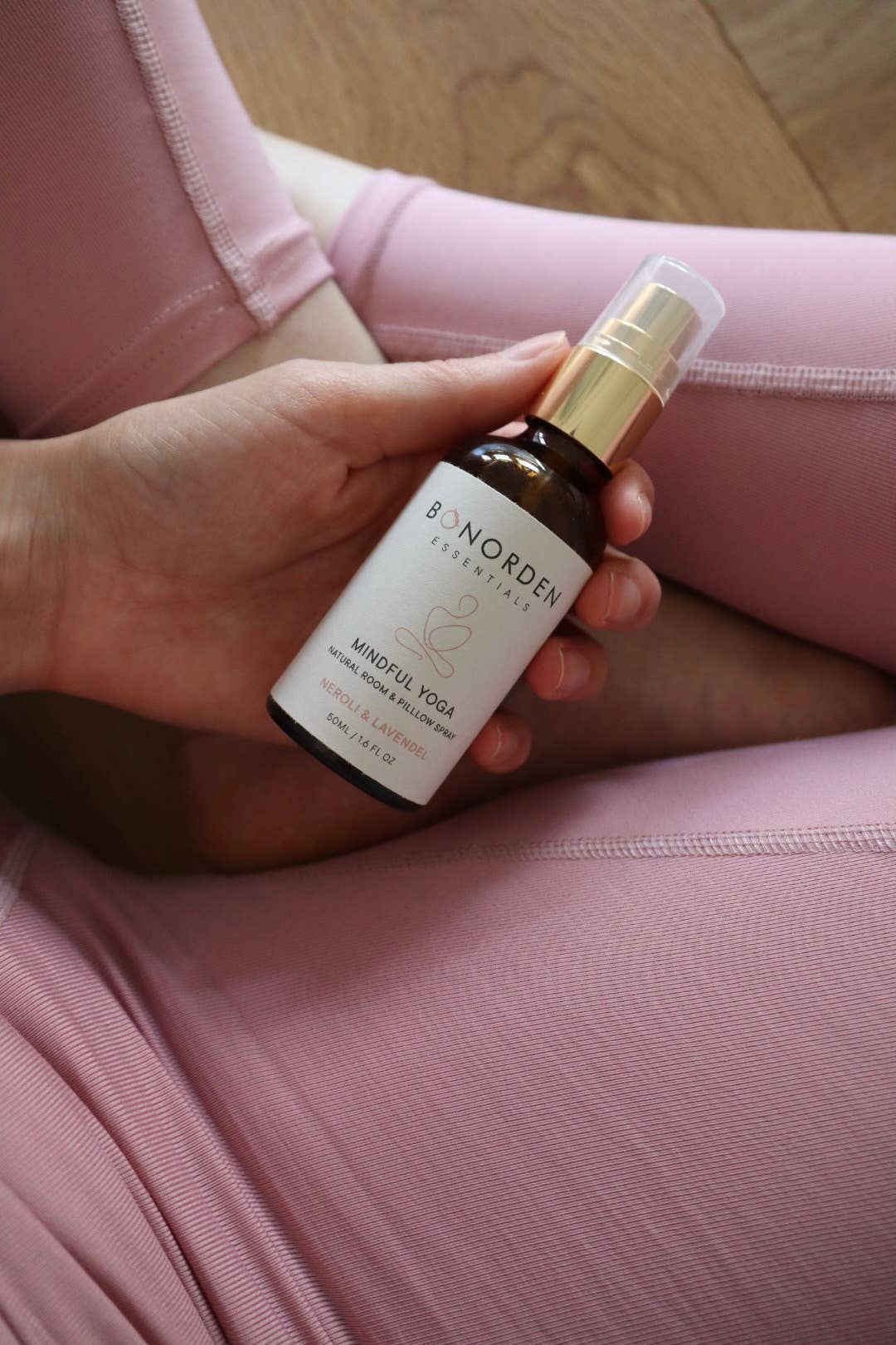 Hand hält eine BONORDEN Mindful Yoga Sprayflasche über rosafarbener Yogahose. Natürliches Raumspray mit Neroli und Lavendel für Ruhe und Achtsamkeit beim Yoga.