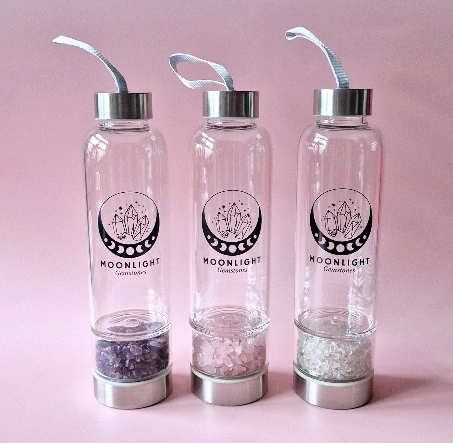 Drei Moonlight Crystal Glas-Trinkflaschen mit unterschiedlichen Edelsteinen im Bodenfach vor rosa Hintergrund.
