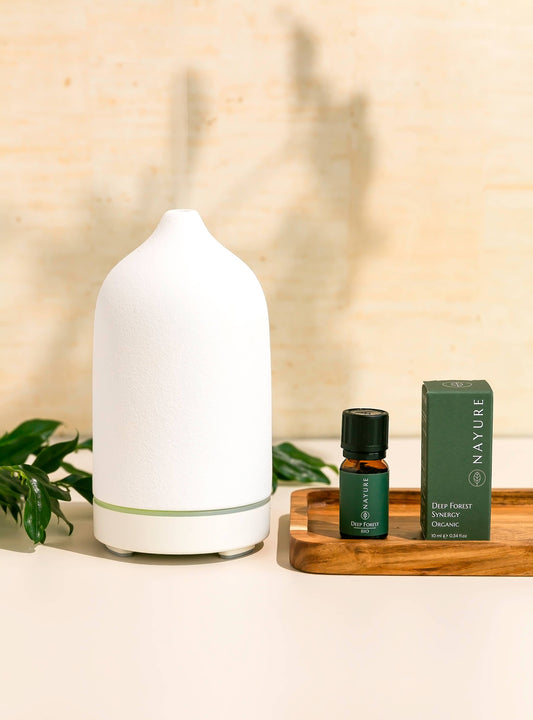 Weißer Keramik-Diffuser mit ätherischem Öl und Verpackung auf einem Holztisch. Natürliche Deko mit Pflanzen im Hintergrund – ideal für Aromatherapie und Entspannung.