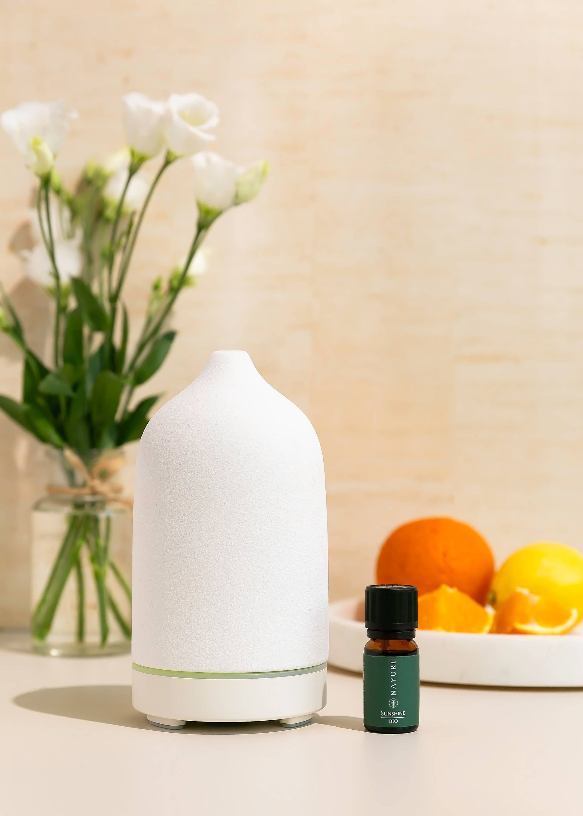 Weißer Aroma-Diffuser mit grünem ätherischem Öl-Fläschchen, arrangiert mit Blumen und Zitrusfrüchten im Hintergrund. Sanftes, helles Ambiente für Wohlbefinden.