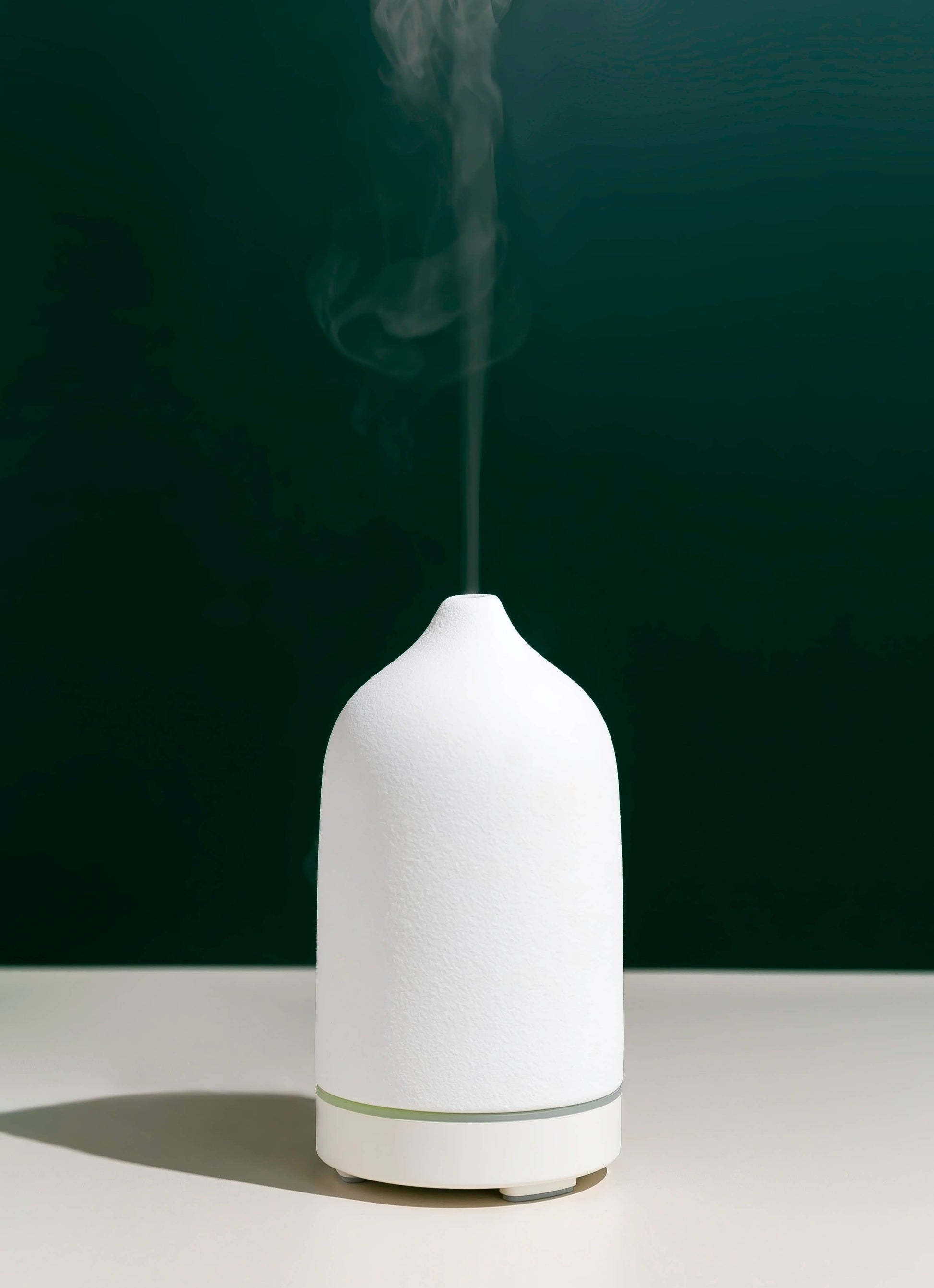 Weißer Keramik-Diffuser mit aufsteigendem Dampf vor grünem Hintergrund. Moderner Raumduft-Vernebler für beruhigende Aromatherapie.