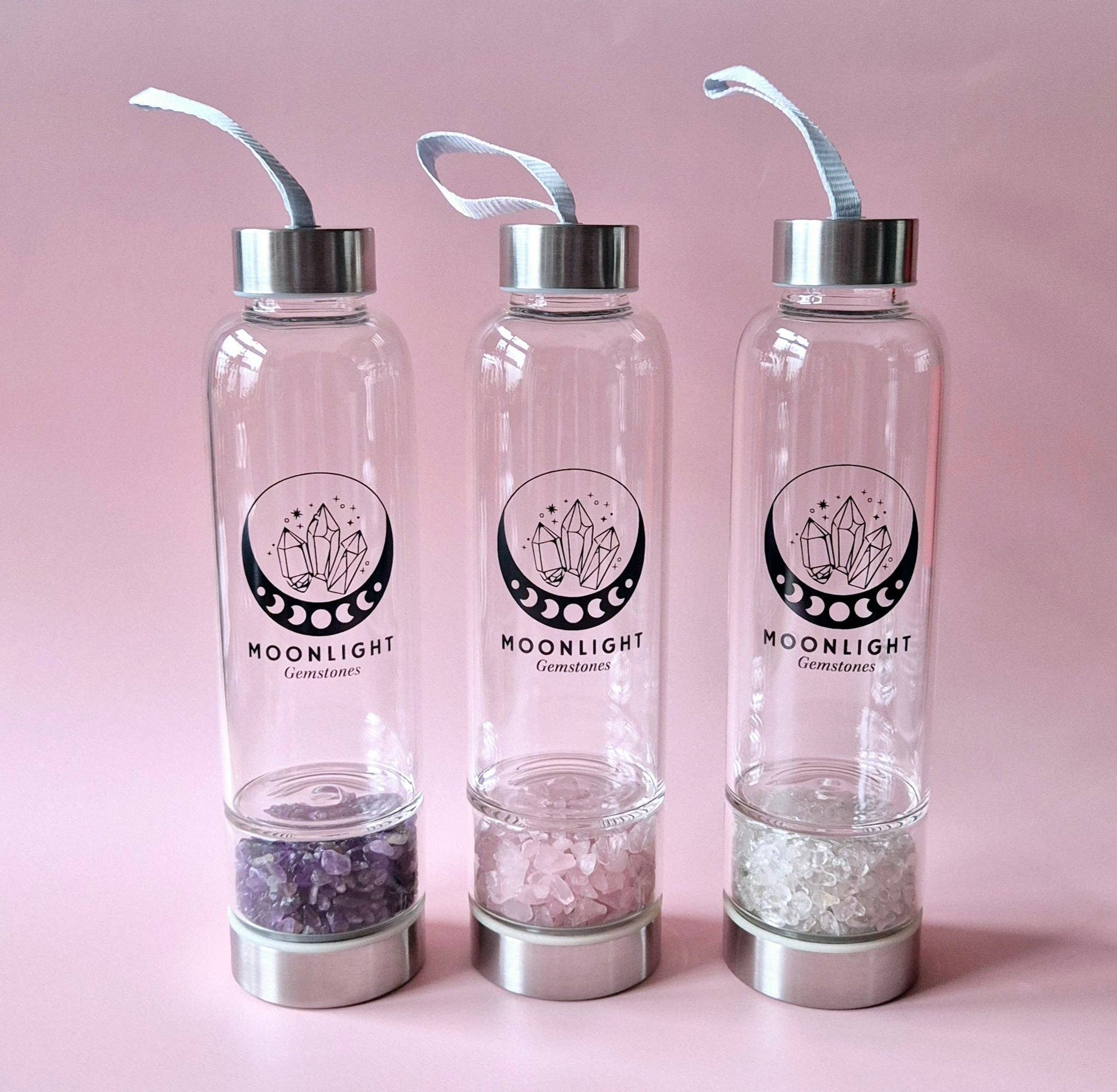 Drei Moonlight Crystal Glas-Trinkflaschen mit unterschiedlichen Edelsteinen im Bodenfach vor rosa Hintergrund.