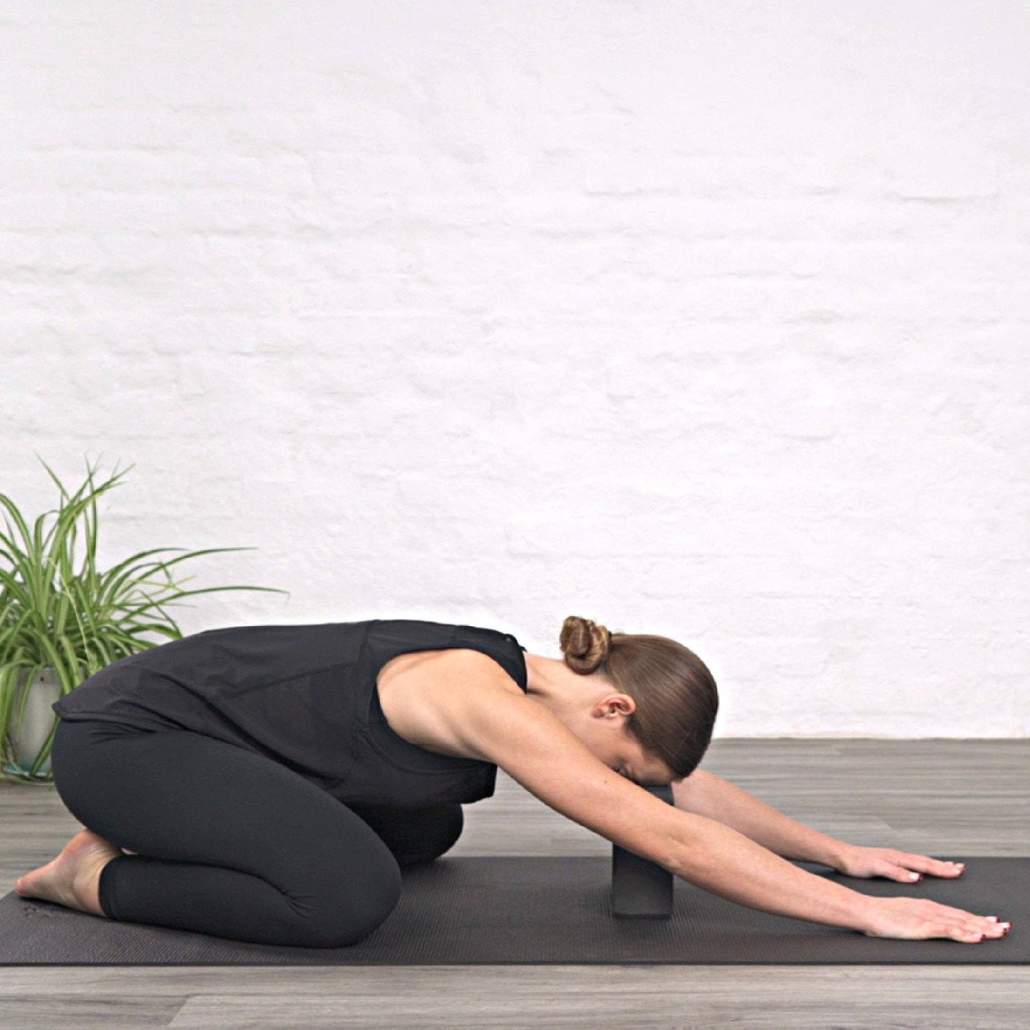 Person in Kinderpose auf schwarzer Yogamatte mit Unterstützung durch einen schwarzen Myga Yogablock unter der Stirn. Entspannende Yoga-Haltung mit Hilfsmittel.
