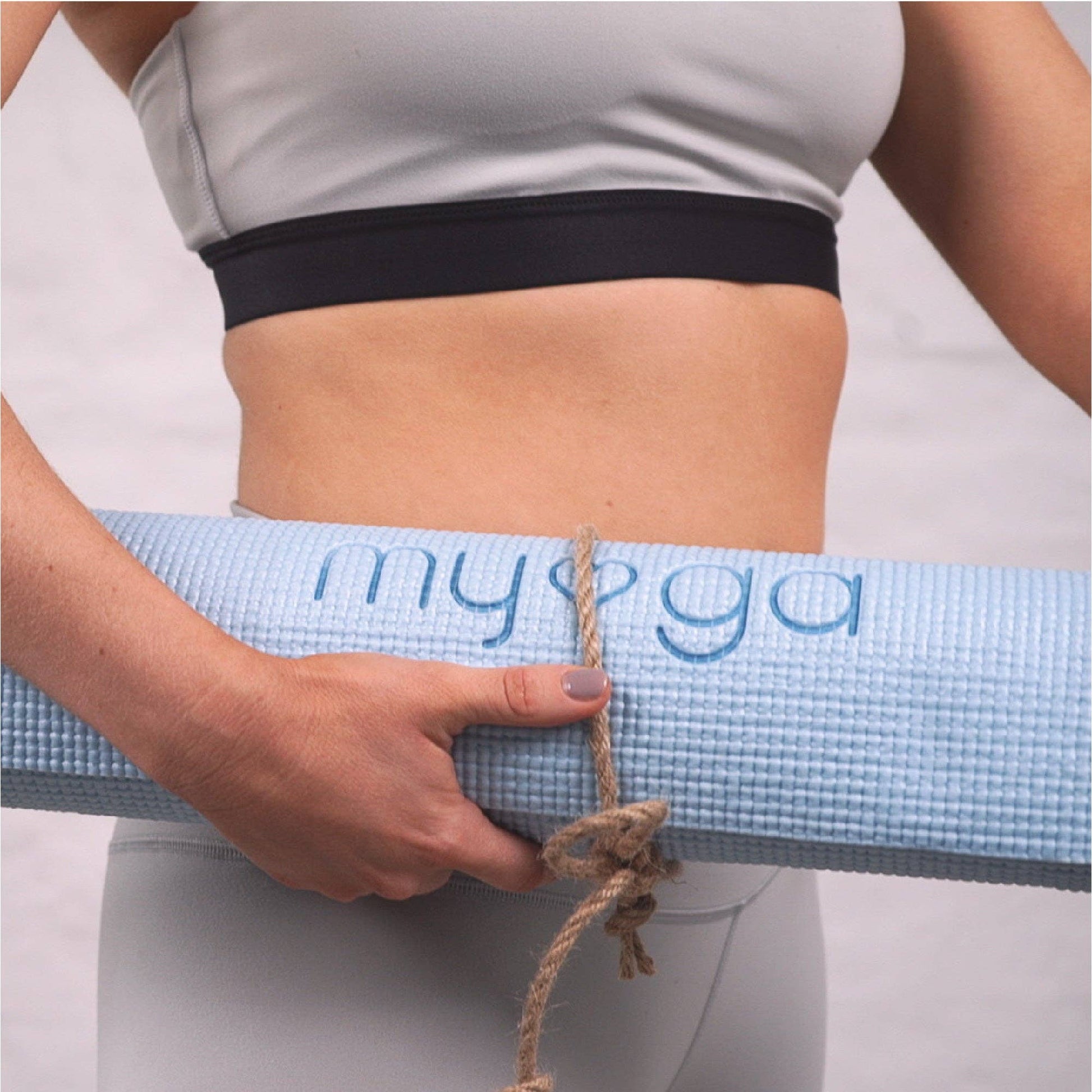 Person in Sportkleidung hält eine zusammengerollte hellblaue Myga Yogamatte mit Trageband in den Händen.